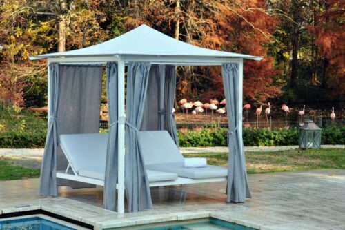 Pavilion mit 4 beams, grey curtains, a white pointy roof next to a pool in a garden surrounded by trees with autumn colours.