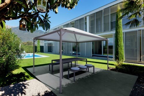 Pavilion with thin bars and a white roof. Covers a table and a bench. Pavilion stands in front of a big building and a garden.