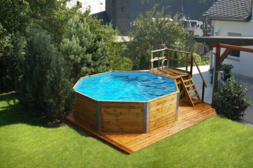 Weka Massivholzpool mit Sandfilteranlage. Ein Pool aus Massivholz steht in einem Garten auf einem Holz boden in der gleichen Farbe. Der Swimmingpool ist gefüllt und hat auch eine Holztreppe mit einer silbernen, metallen Leiter für den Einstieg ins Wasser. Pool steht vor einem weissen Haus.
