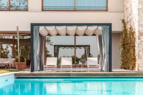 Der Unosider Pavillon Mood YPE vor einer grossen Villa mit Pool mit weissen Liegestühlen, Vorhängen und Dach